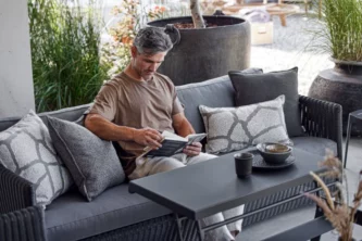 Man in taupe t-shirt reading on a modern grey upholstered sofa with an adjustable black coffee table on a patio