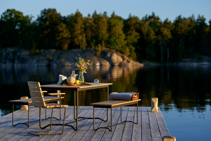 Square outdoor dining table with teak slat top and black metal frame with coordinating benches and armchair on a lake pier