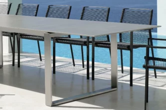 Modern patio dining table with steel base and thin ceramic top surrounded by angular black rope chairs near the ocean