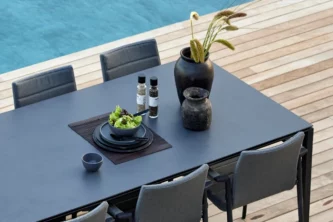 Overhead view of rectangular outdoor dining table with a black ceramic tabletop with a salad bowl and plates on a light wooden deck near a pool