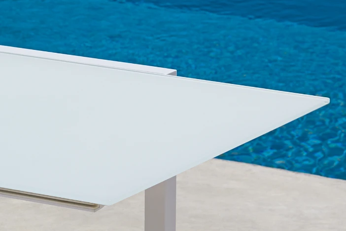 Edge of a rectangular white tempered glass patio dining table next to a bright blue swimming pool