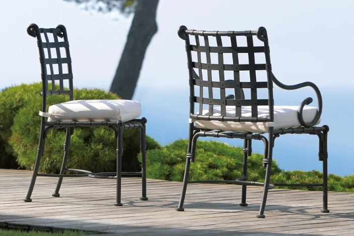 Two Tuscan-style outdoor dining chairs with black wrought metal frames and lattice backrests with white seat cushions on a deck overlooking water