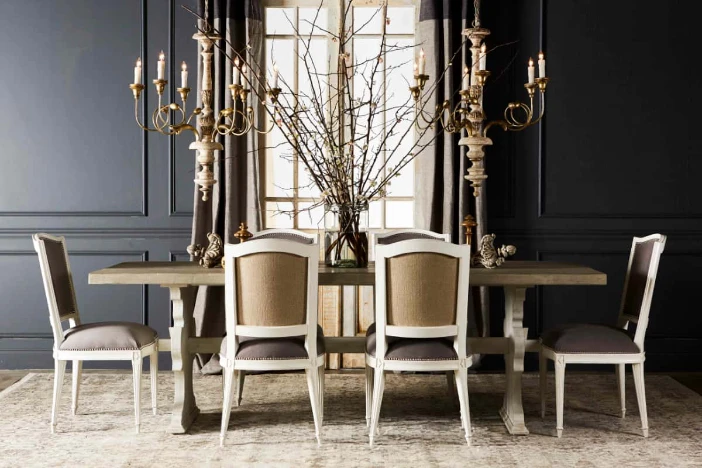 Dining set with rectangular wood table, white shabby chic side chairs with taupe upholstery below two distressed brass and wood spindle chandeliers