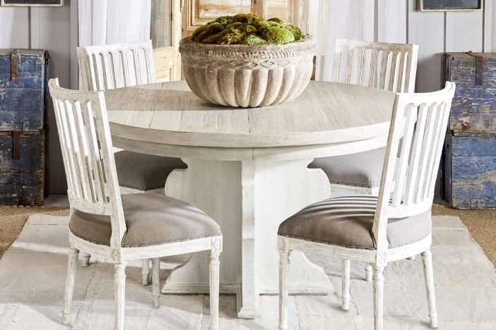 Round wooden dinette table with decorative bowl surrounded by four distressed white side chairs with taupe seat upholstery