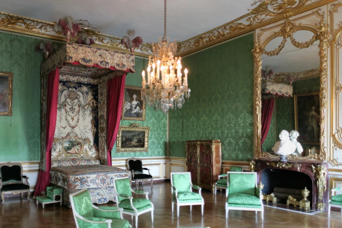 Bedroom in the Palace of Versailles with light green wallpaper and coordinating upholstered armchairs in front of an ornate red curtained bed