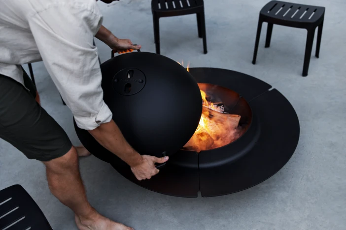 Man placing a protective spark guard cover over a the flames of a wood-burning fire pit on a concrete patio