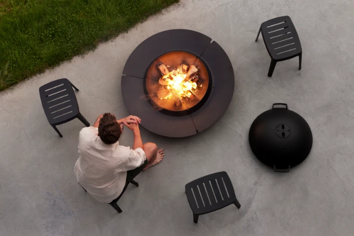 Overhead view of round wood-burning fire pit and spark guard on a concrete patio with well-spaced black stools and a seated man