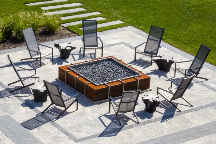 Modern black Adirondack chairs arranged around a square oxidized metal gas fire pit on a tiled backyard patio