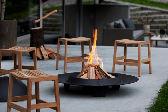 Black steel wood-burning fire pit on cement patio with low wooden stools