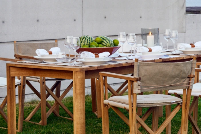 Teak outdoor dining table and folding armchairs with light brown canvas seats and backs on a lawn