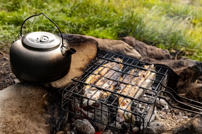 Fish being grilled over a stone fire pit with charcoal and a kettle nearby