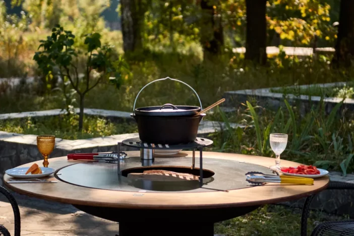 Round fire pit table with a kettle simmering on a grill above the central wood-burning opening and two place settings