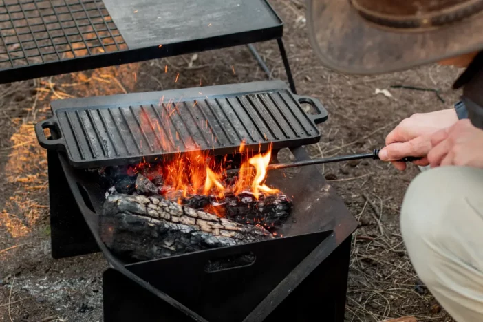 Man stoking wood fire in a black metal collapsible fire pit with griddle and grate