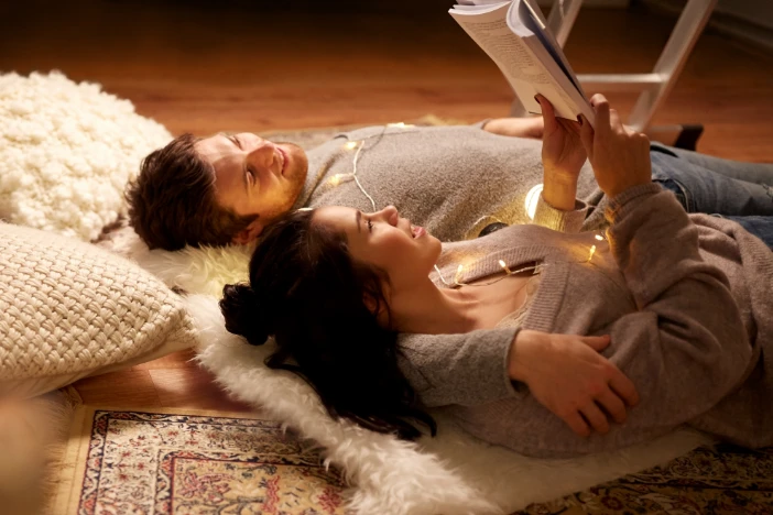 Couple enjoying a book while laying on a white furry rug with textured pillows