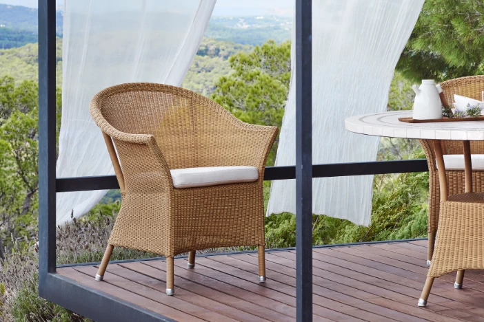 Light brown wicker armchair on a wooden deck with white curtains blowing in the wind