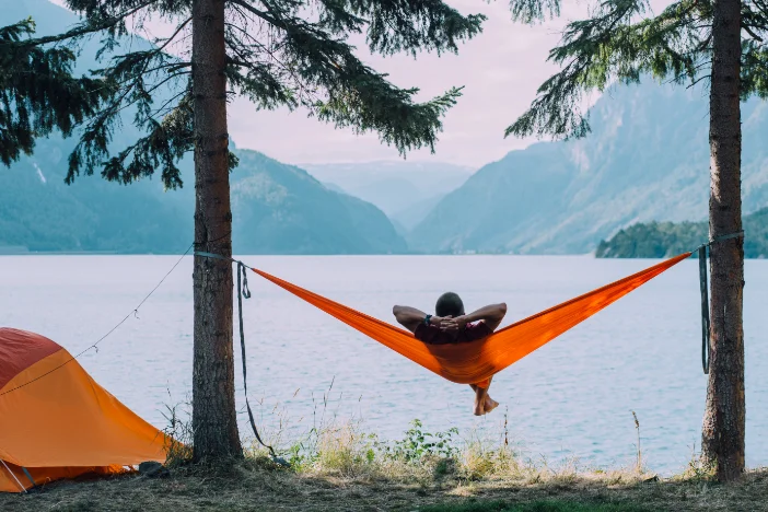 Man laying in an orange hammock strung between trees on a lake next to a tangerine camping tent