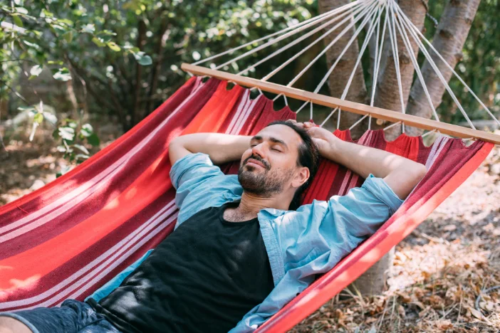 Bearded man in blue laying on a red and orange striped bohemian-style hammock hanging between trees