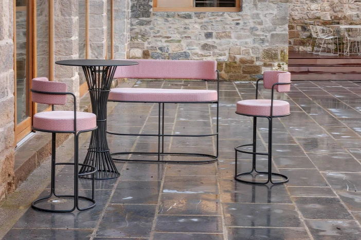 Two dark grey framed bar stools and loveseat with rounded pink seat and back cushions around a bar table outside a stone house