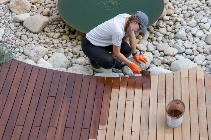 Overhead view of a woman in a t-shirt and baseball cap staining a hardwood deck next to a rocky area