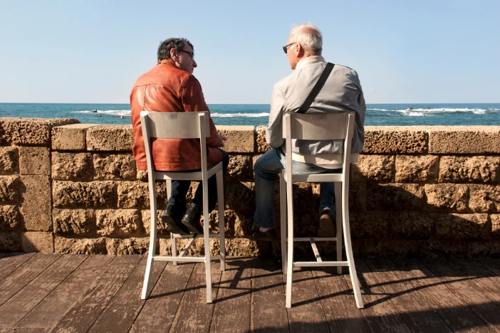 Two older men sit on tall barstools behind a stone wall overlooking the water on a clear day