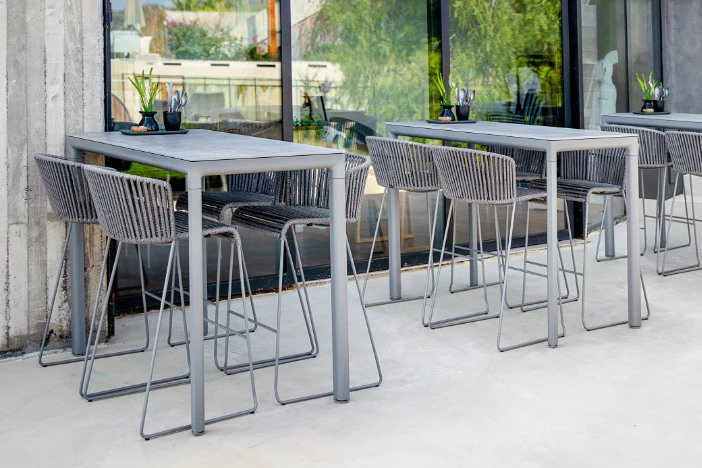 Light grey wireframe bar chairs with soft rope seat and backrests tucked under matching bar tables on patio