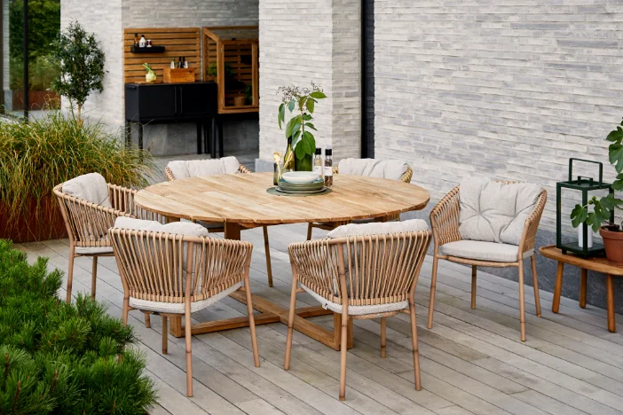 Round wooden patio dining table and rattan chairs with beige cushions on a hardwood deck