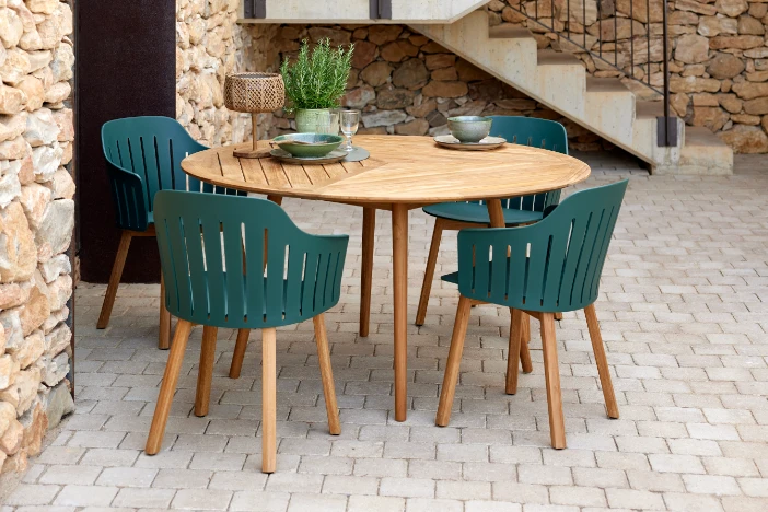 Round teak dinette surrounded by modern chairs with dark green plastic bodies on a brick patio