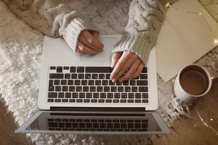 Woman in heavy sweater types at a laptop with a cup of coffee during winter