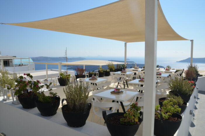 Large natural colored rectangular shade sails stretch over white dining sets overlooking a coast