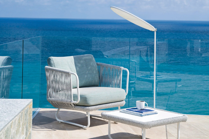 Modern white outdoor lounge chair with rope body, teal cushions, matching table, and personal shade on a glass‑walled patio overlooking the water