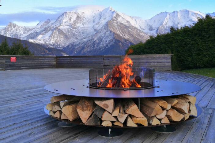 Round steel AK47 fire pit with blazing fire and log storage on a deck with a mountain view