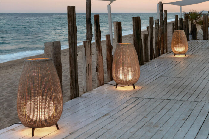 Three large light brown rattan Bover outdoor floor lamps on a weathered beachfront deck