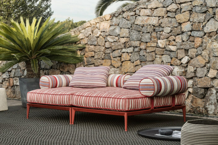Crimson metal‑framed two‑piece outdoor daybed with striped cushions and bolster pillows on a grey rug by a stone wall