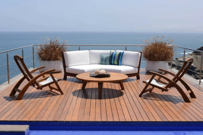 Two curving teak folding lounge chairs with sling seats and a curved loveseat with white cushions around a coffee table on an elevated pool deck with ocean view