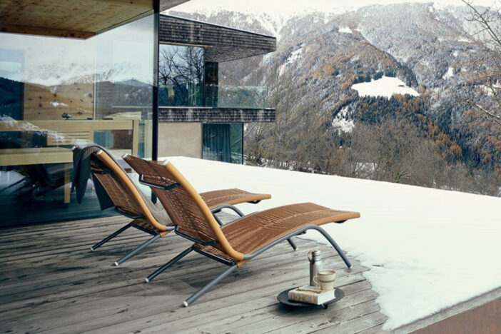 Two modern reclining sunloungers with grey metal frames and tan woven bodies on a snow‑covered deck with a mountain view