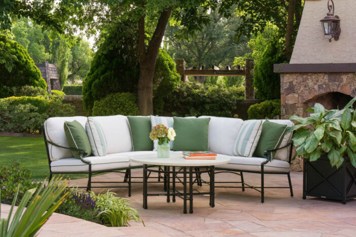 Green metal‑framed corner sectional with white cushions and accent pillows on a stone patio with lush greenery