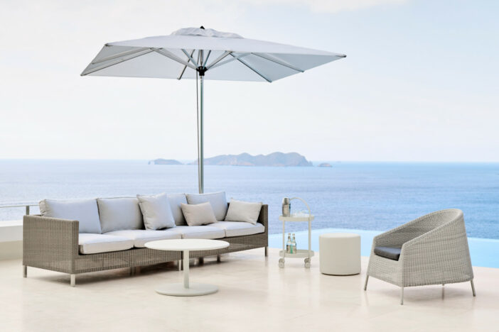 White‑grey and beige outdoor wicker lounge chair and sofa with accent tables under a square umbrella on a seaside patio