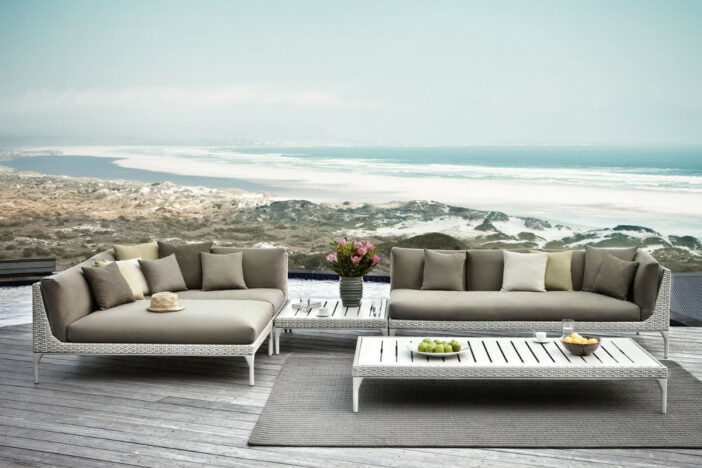 Low white outdoor wicker sectional with brown cushions and pillows on a weathered deck overlooking a rocky coastline