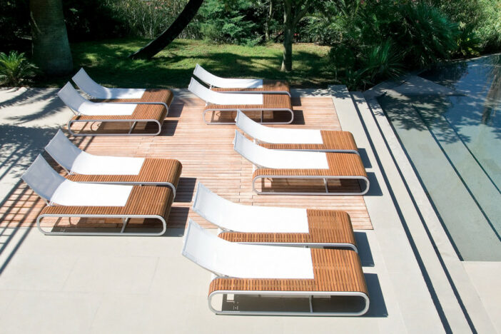 Ten chic sunloungers with curved white sling backs and teak slat bodies arranged by a large step‑down pool