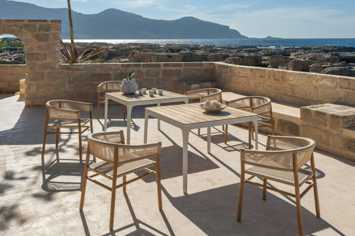 Light wood outdoor dining chairs with wraparound woven backs and seats around square tables on a stone‑walled Mediterranean terrace