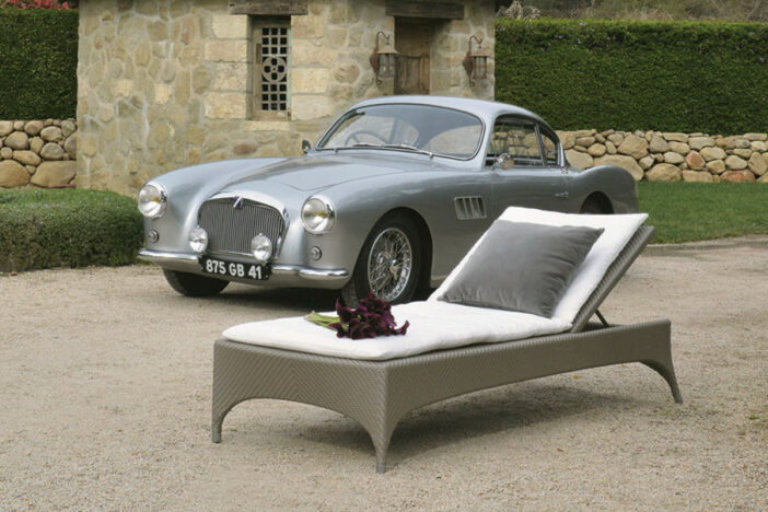 Streamlined gunmetal grey woven sunlounger with thin white cushion beside a vintage silver sports car on a gravel drive by a stone wall