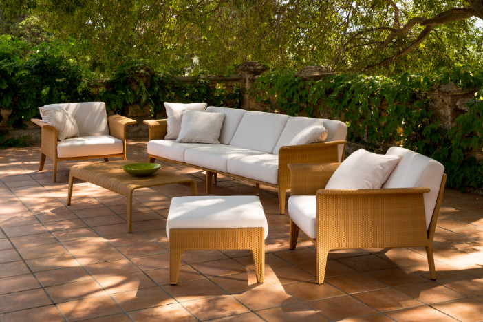 Upscale outdoor wicker lounge set with golden brown tight weave, flared arms, and plush white cushions on a tiled patio surrounded by trees