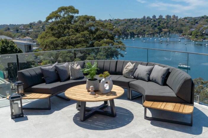 Semi‑circular outdoor sectional sofa with built‑in teak side tables and angular dark grey cushions on an elevated marble deck above a marina