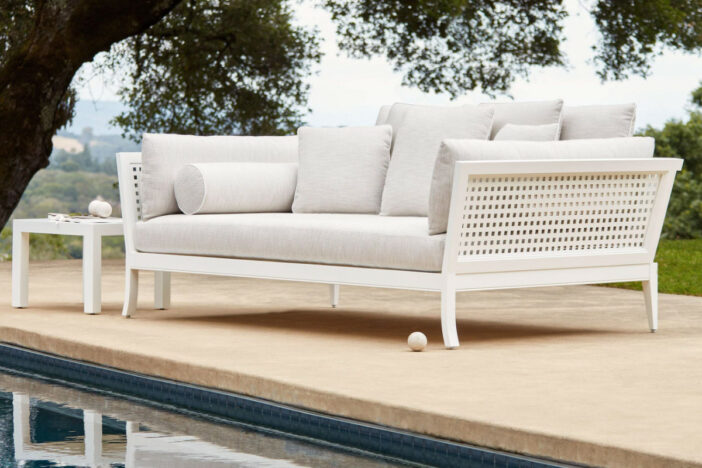 Distinguished white wooden loveseat with trellis sides and deep off‑white cushions and pillows beside a pool under a tree