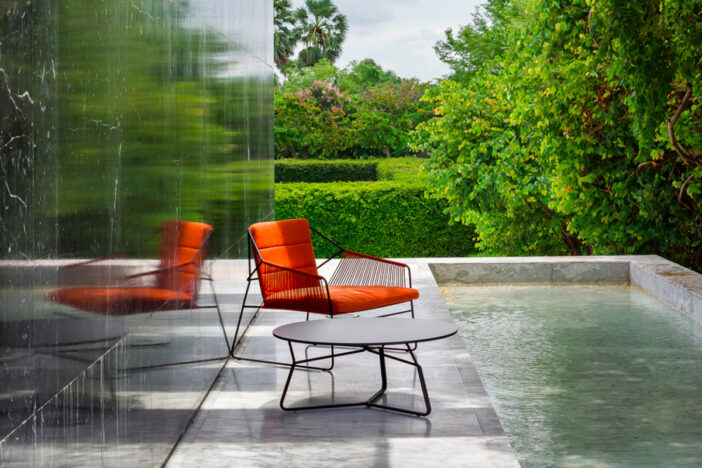 Ultra‑contemporary outdoor accent chair with vibrant orange cushions and roped sides on a narrow marble strip by a decorative pool