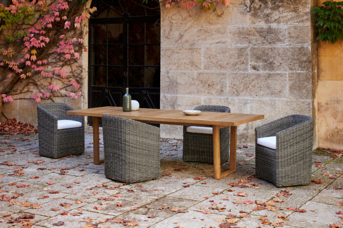 Grey outdoor wicker dining chairs with rounded contours and angular arms around a rectangular teak dining table on a leaf‑strewn stone patio