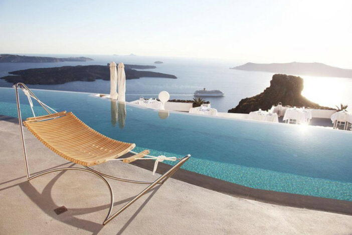 Minimalist curved wood slat hammock supported by a dynamic stainless steel tube frame beside an infinity pool overlooking the sea