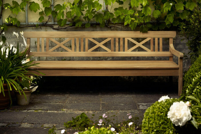 Wide traditional teak bench with cross‑slat back on a concrete garden patio with climbing vines