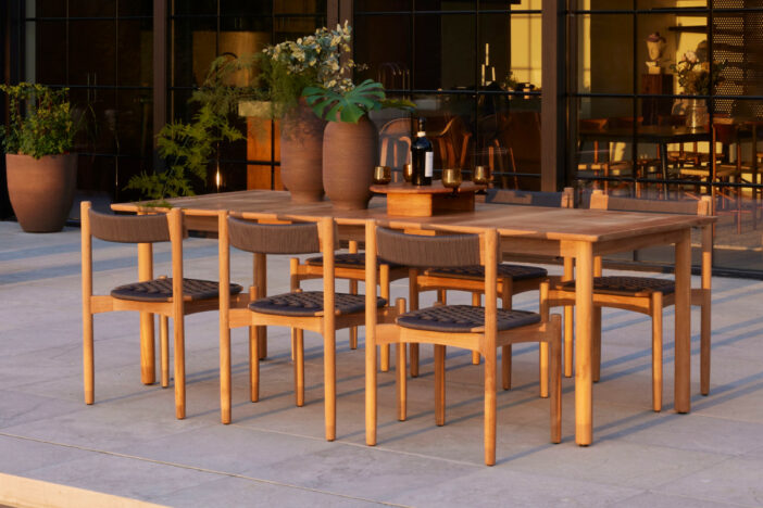 Classic teak dining set with dark grey woven seats and wrapped backs around a rectangular table on a marble patio at sunset