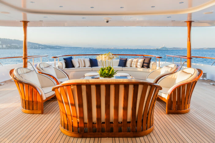 Glossy marine‑finished teak lounge seating with wide vertical slat wraparound design and white cushions on a covered yacht deck